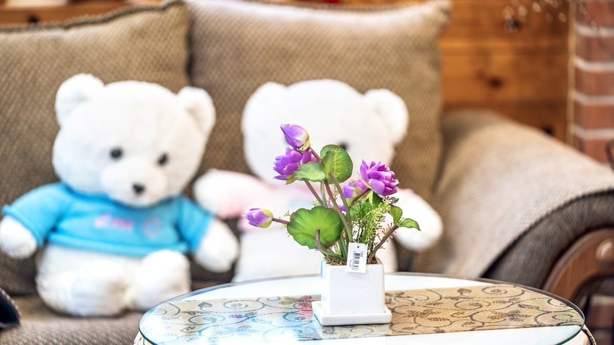 A white teddy bear in a blue shirt on a couch, a white teddy bear on a cushion, and a small white vase with purple flowers on a table.