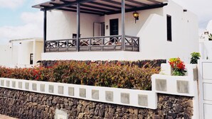A white building with a covered porch, stone wall, and a small garden with colorful flowers. - CASA VINCA, quiet holiday home near the beach (Puerto del Carmen)