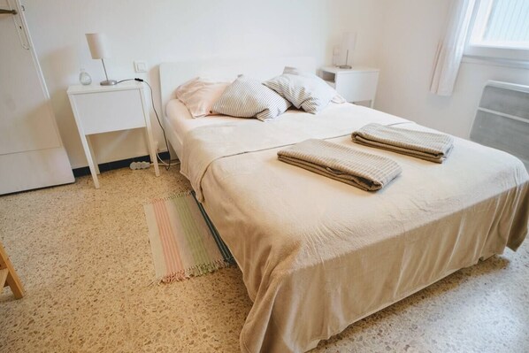 A neatly made bed with pillows, a bedside table with a lamp, and a small rug on a speckled floor.