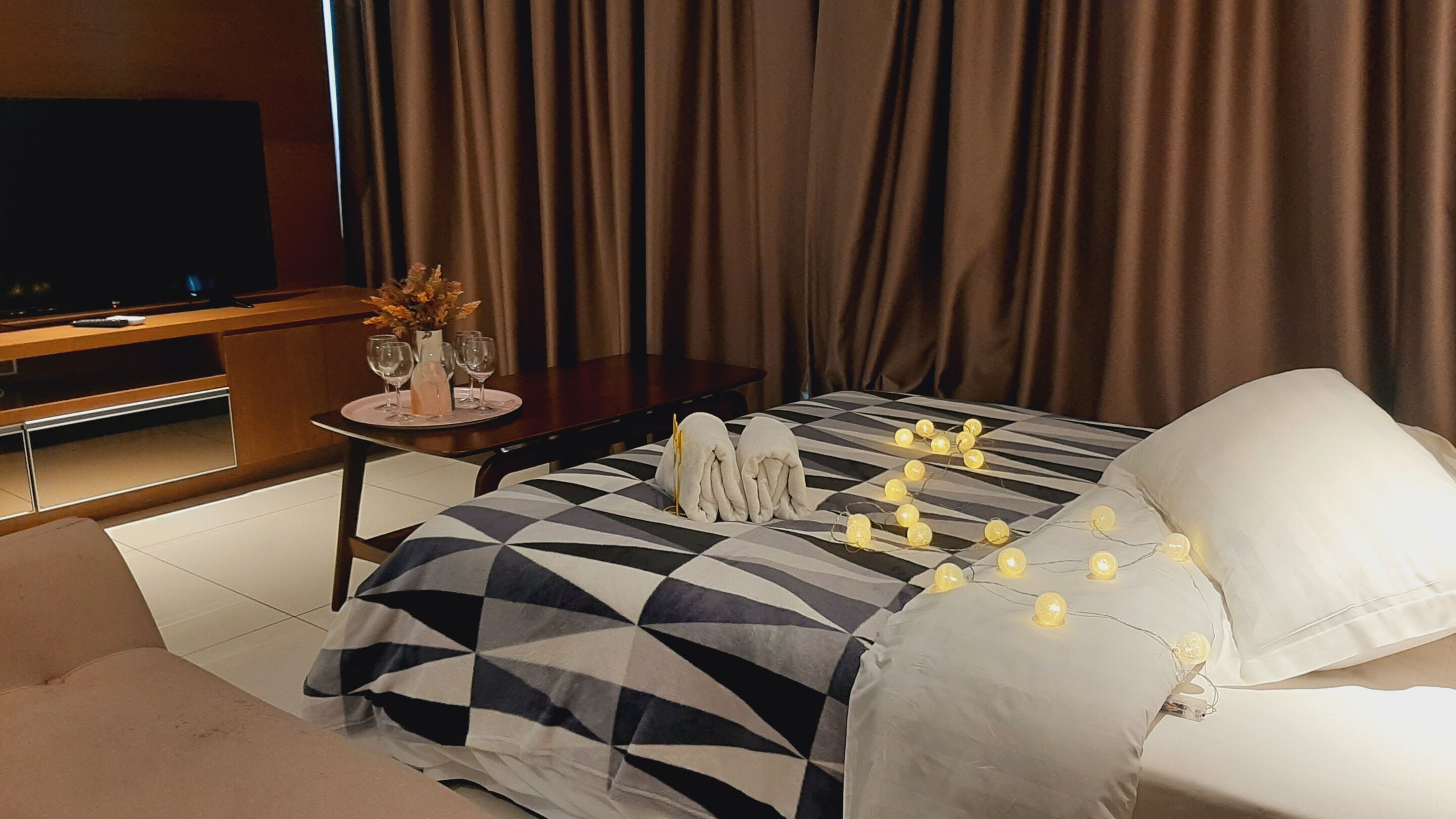 Una habitación de hotel moderna con una cama grande, un televisor, una mesita con un florero y vasos, y cortinas.
