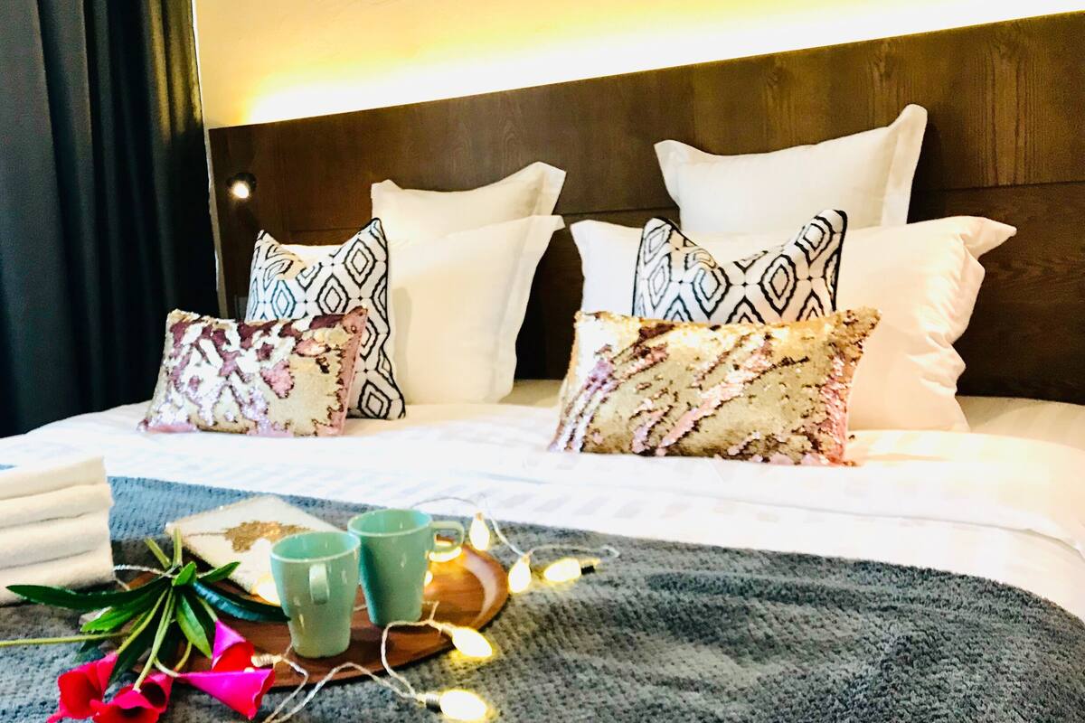 Ein Hotelzimmer mit einem ordentlich bezogenen Bett, gemusterten Kissen, einem Nachttisch mit Lampe und einem Fenster mit Vorhängen.