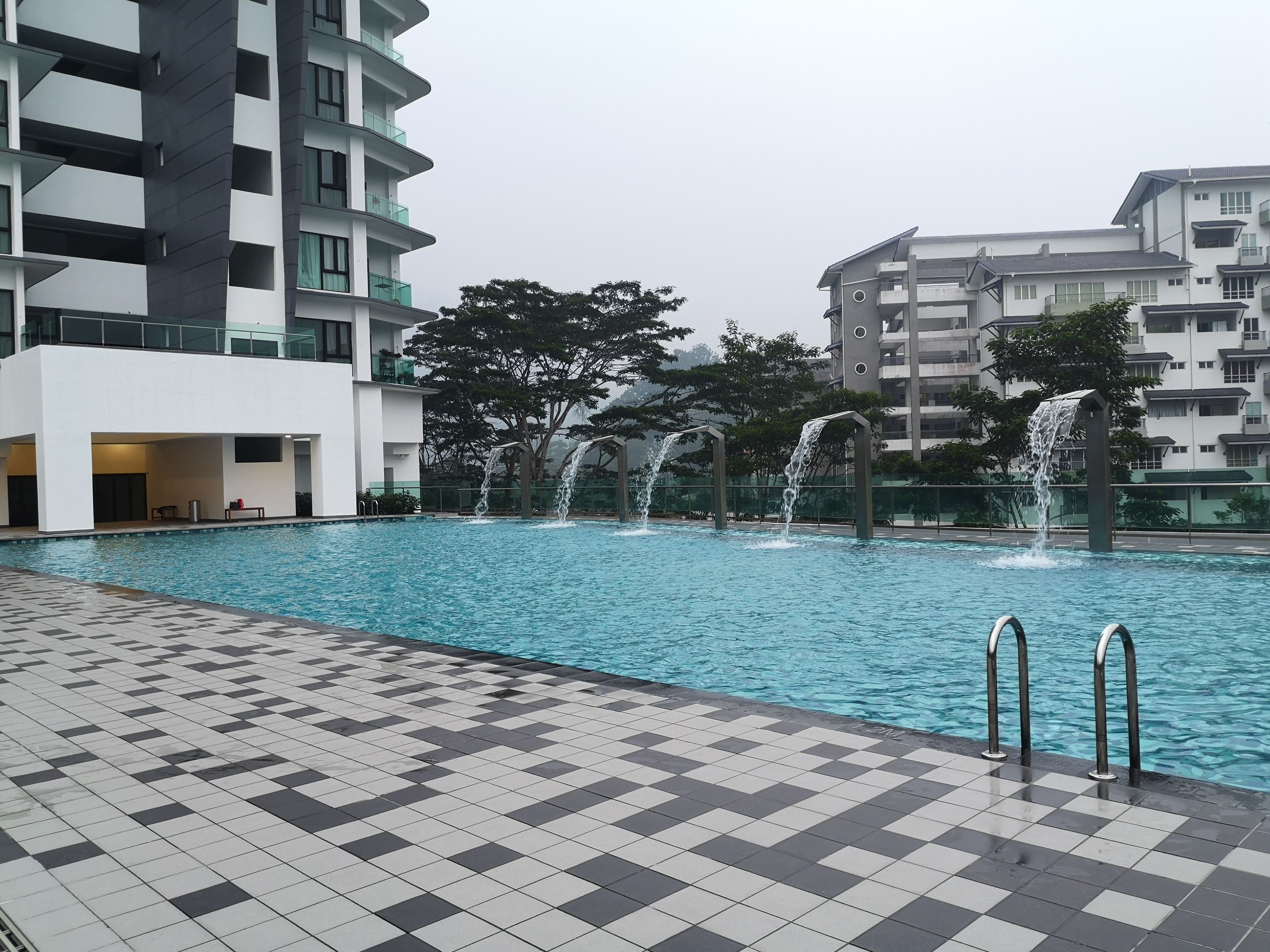 A swimming pool with a checkered tiled deck, surrounded by modern buildings with balconies and a waterfall feature.