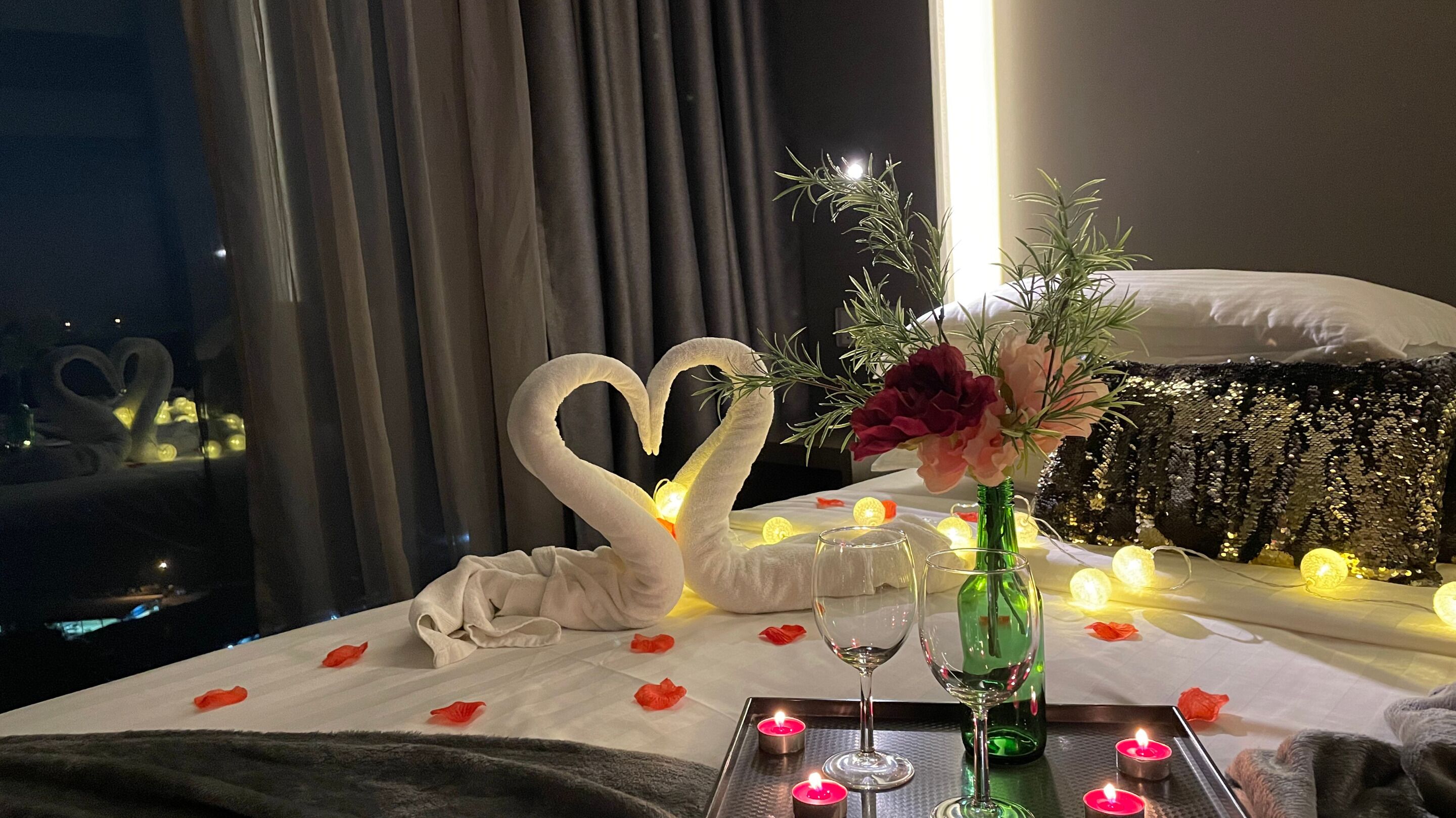 Un lit agrémenté d’une décoration en forme de cygne en forme de cœur, un vase rempli de fleurs, des bougies et un verre de vin posé sur un plateau.