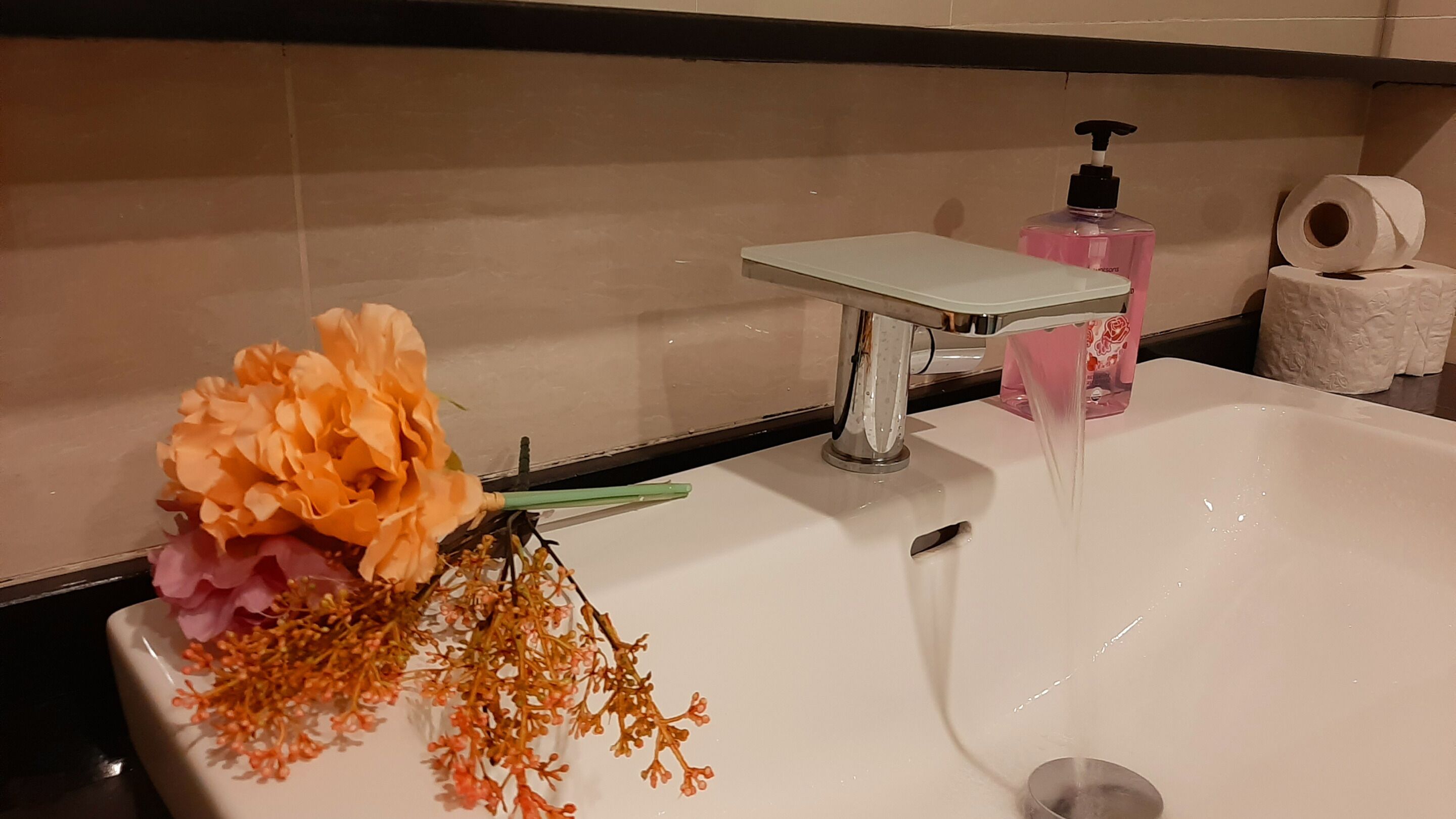 Un lavamanos con grifo, un arreglo floral, un dispensador de jabón y un rollo de papel higiénico.