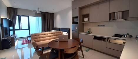 A modern hotel room with a sofa, dining table, chairs, a laptop, a kitchen area, and a large window.