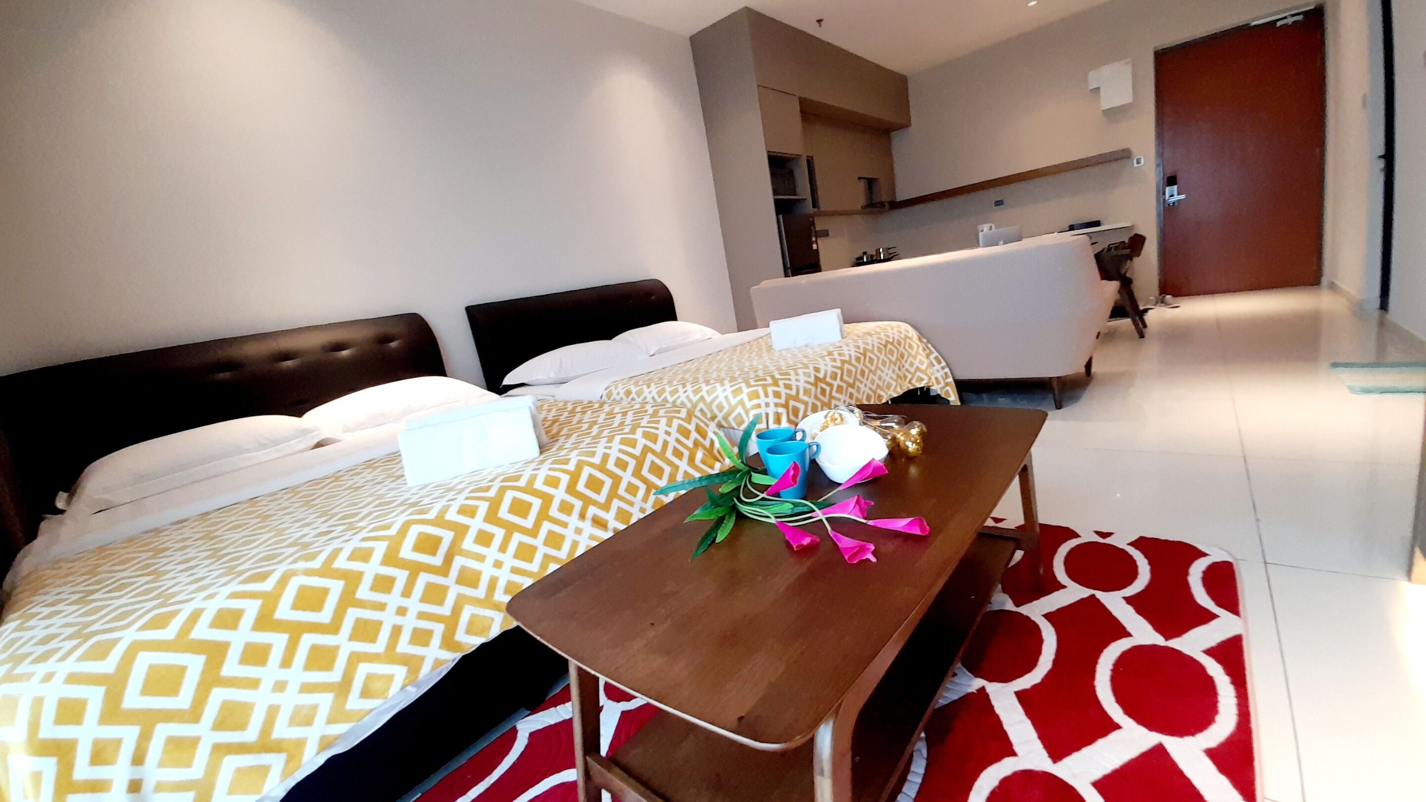 A hotel room with two beds, a wooden table with a bouquet, and a red patterned rug.