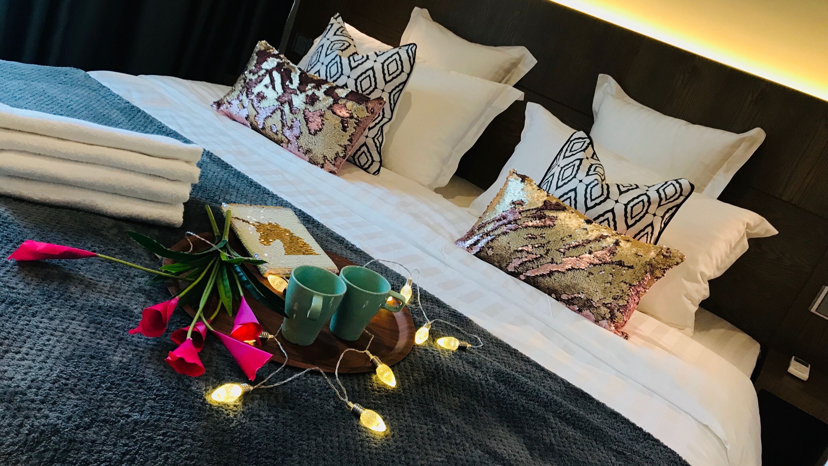 Un lit bien fait, agrémenté d’oreillers à motifs, une table de chevet avec deux tasses vertes et une guirlande de petites lumières chaleureuses.