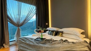 Un dormitorio con un ventanal grande, una cama con almohadas blancas y una almohada decorativa, una mesita de noche con un florero y copas de vino, y vistas a un paisaje montañoso.