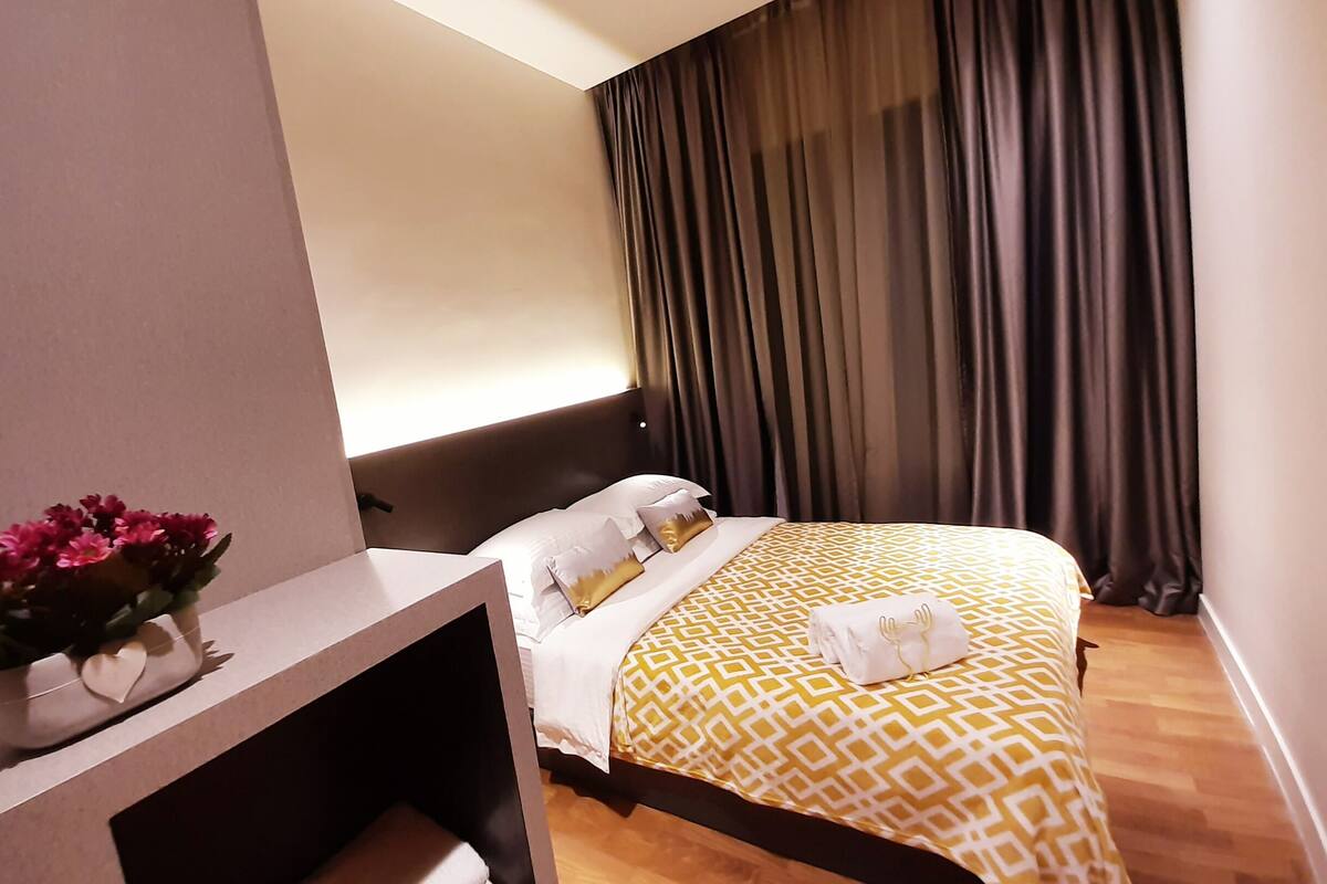 Ein Hotelzimmer mit einem Bett, einem Nachttisch mit Blumenvase und einem großen Fenster mit Vorhängen.