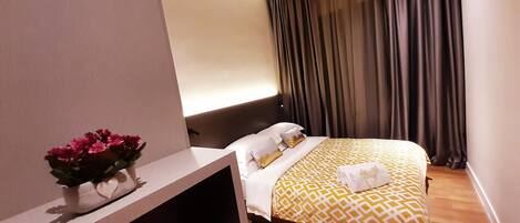 A hotel room with a bed, a bedside table with a vase of flowers, and a large window with curtains.