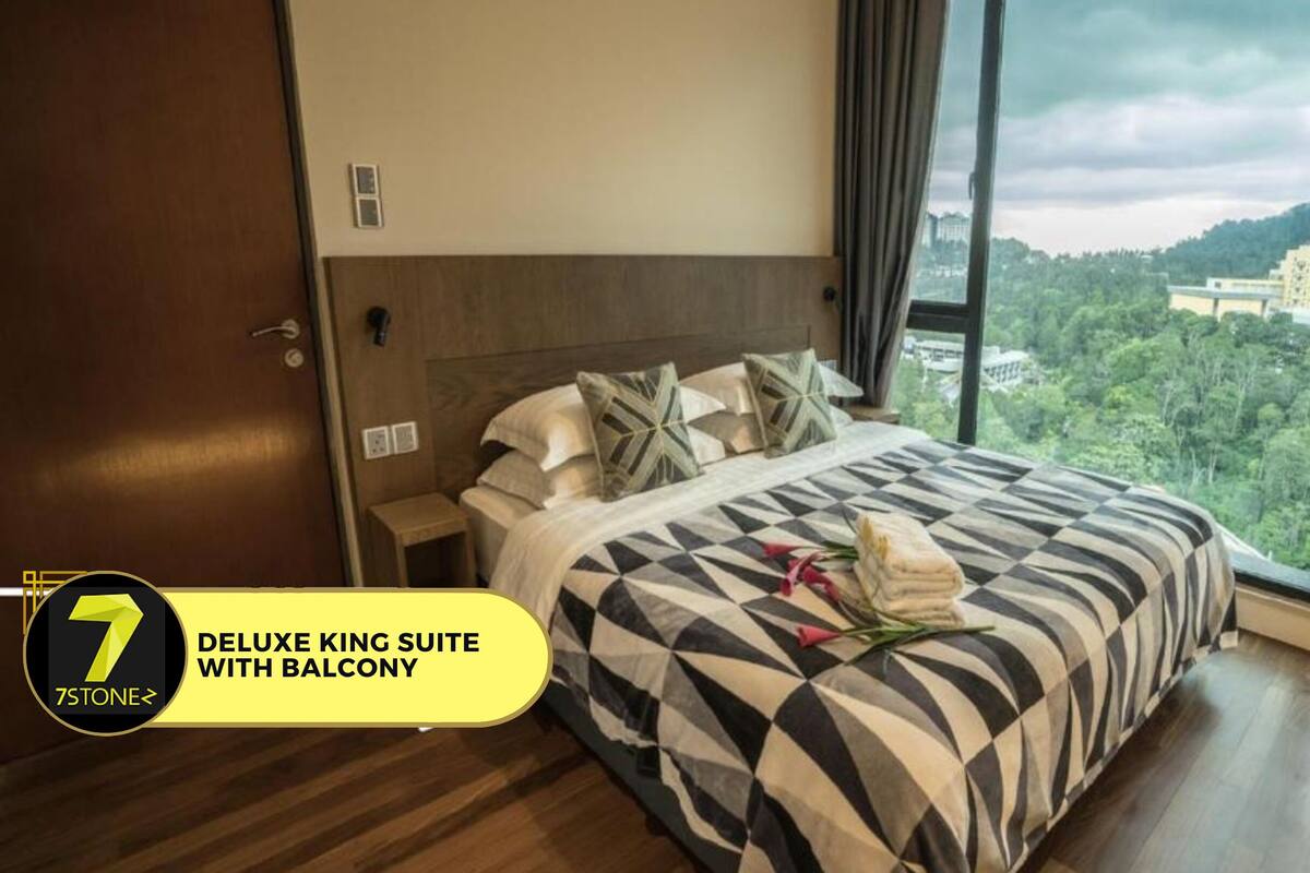 Eine Deluxe-Königssuite mit Balkon, mit einem großen Bett mit gemusterten Bettwäsche, einem Nachttisch und Blick auf eine Stadtlandschaft durch große Fenster.
