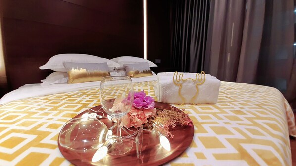 Una cama bien hecha con una colcha estampada, una mesita de noche con un vaso y flores, y una toalla blanca con detalles dorados.