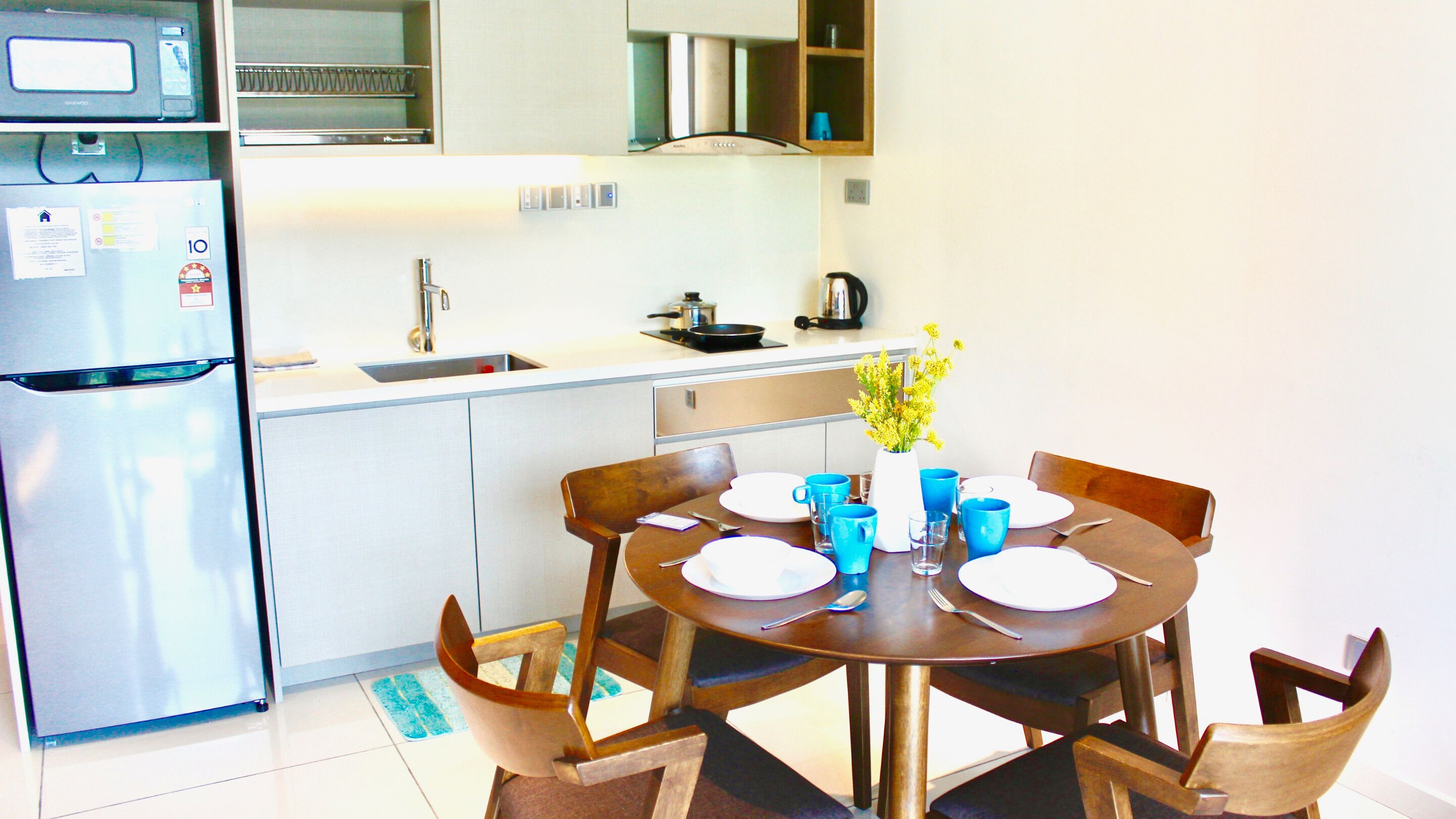 A compact kitchen with a refrigerator, microwave, sink, and stove. There is a dining table with blue cups and plates, and a vase with yellow flowers.
