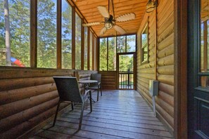 A wooden cabin with a porch featuring a ceiling fan, a table with chairs, and large windows offering a view of the surrounding forest. - Jolly Roger Cabin | Pirate Ship Playground | Luxury Broken Bow Retreat (BROKEN BOW)
