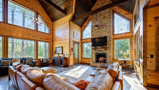A spacious living room with leather sofas, a fireplace, and large windows offering a forest view. - Jolly Roger Cabin | Pirate Ship Playground | Luxury Broken Bow Retreat (BROKEN BOW)