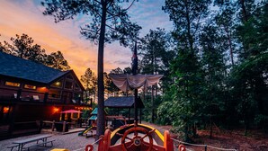 A wooden deck with a ship's wheel, surrounded by trees and a lodge in the background. - Jolly Roger Cabin | Pirate Ship Playground | Luxury Broken Bow Retreat (BROKEN BOW)