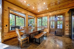 A wooden cabin interior with a large dining table, benches, and large windows offering views of the surrounding forest. - Jolly Roger Cabin | Pirate Ship Playground | Luxury Broken Bow Retreat (BROKEN BOW)