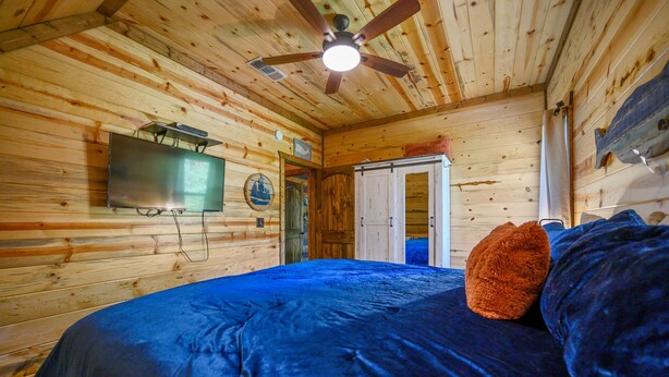 Une chambre dans une cabane en bois, avec un lit, un ventilateur de plafond et un téléviseur fixé au mur.