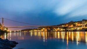 Eine Hängebrücke im Dämmerlicht, mit Lichtern, die sich im Wasser spiegeln.