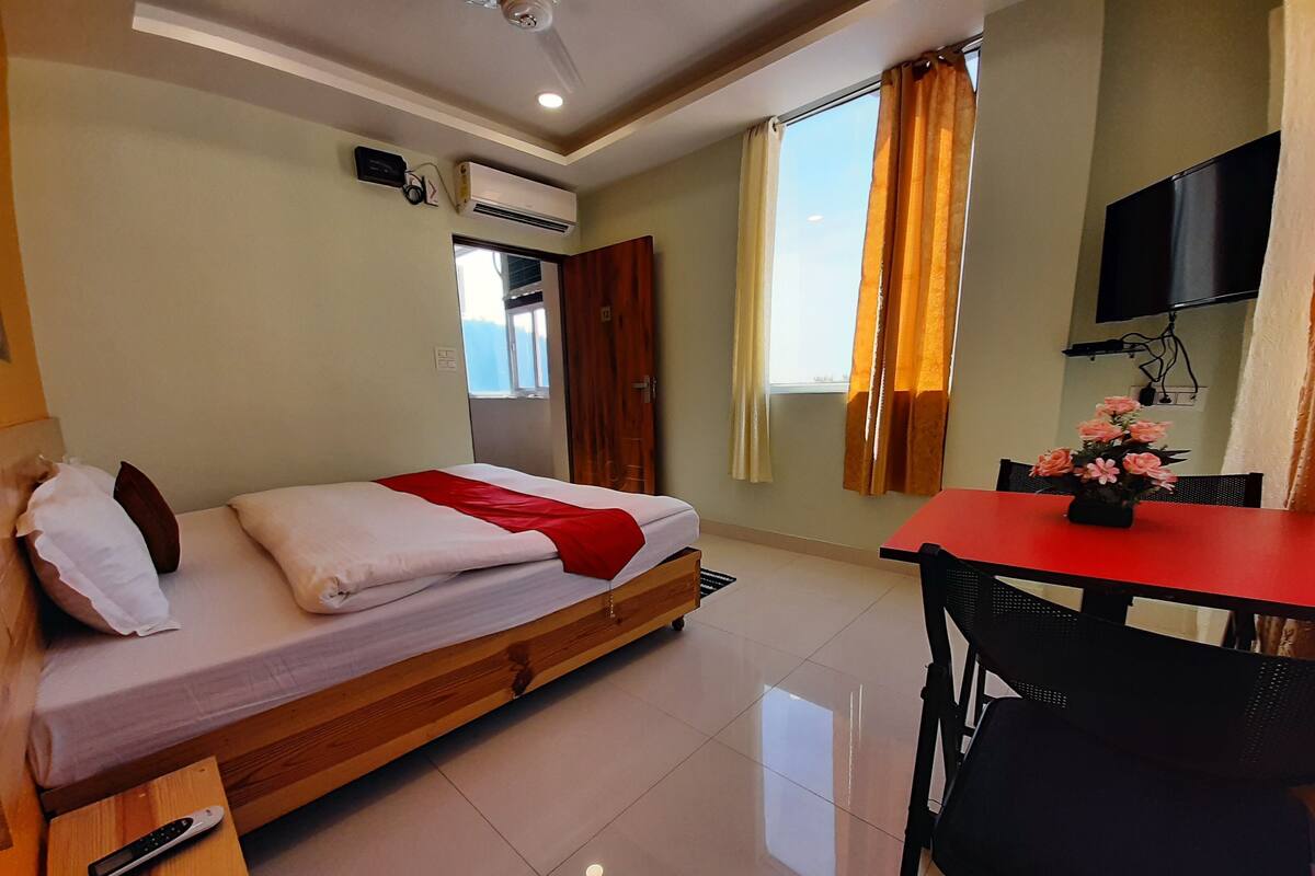 A hotel room with a bed, a desk, a chair, an air conditioning unit, a TV, and a window with curtains.