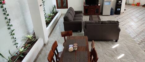A living room with a sofa, a TV, and a dining table with chairs. There is a small garden area with plants and a window with curtains.