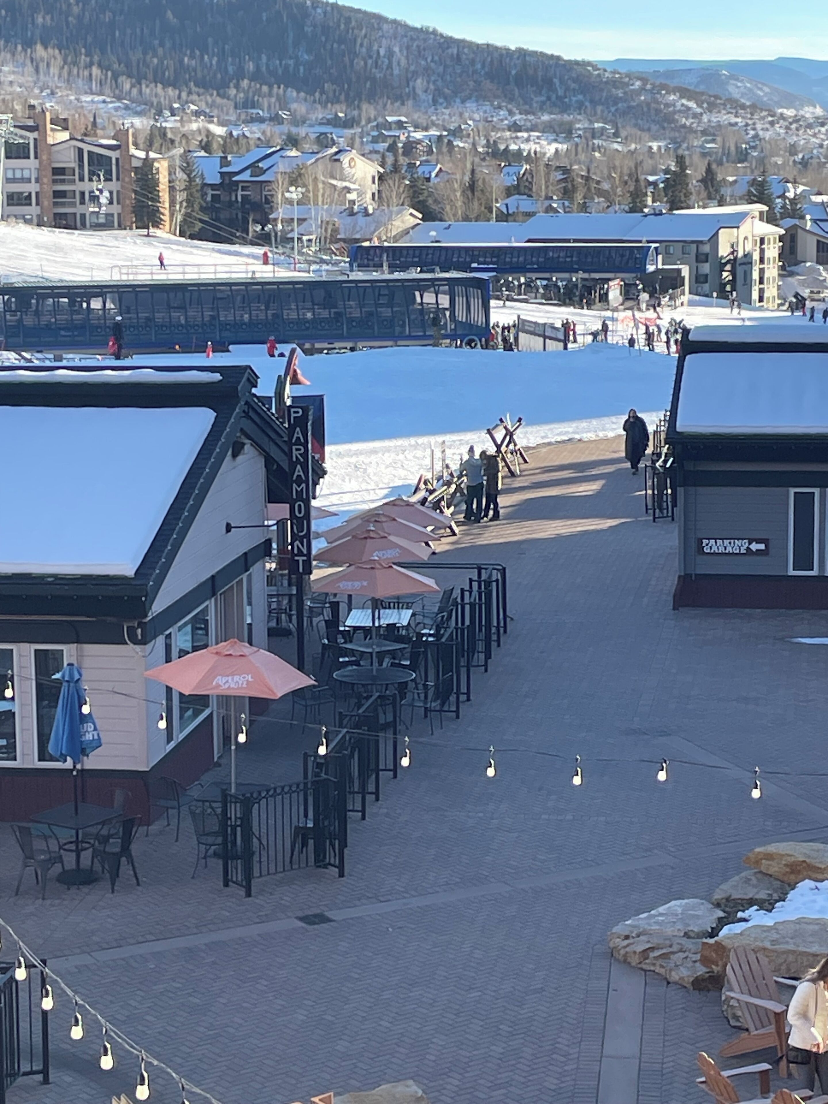 A snow-covered outdoor seating area with tables and chairs, a building with a sign reading "PARADISE", and a view of a town with buildings and snow-covered hills in the background.