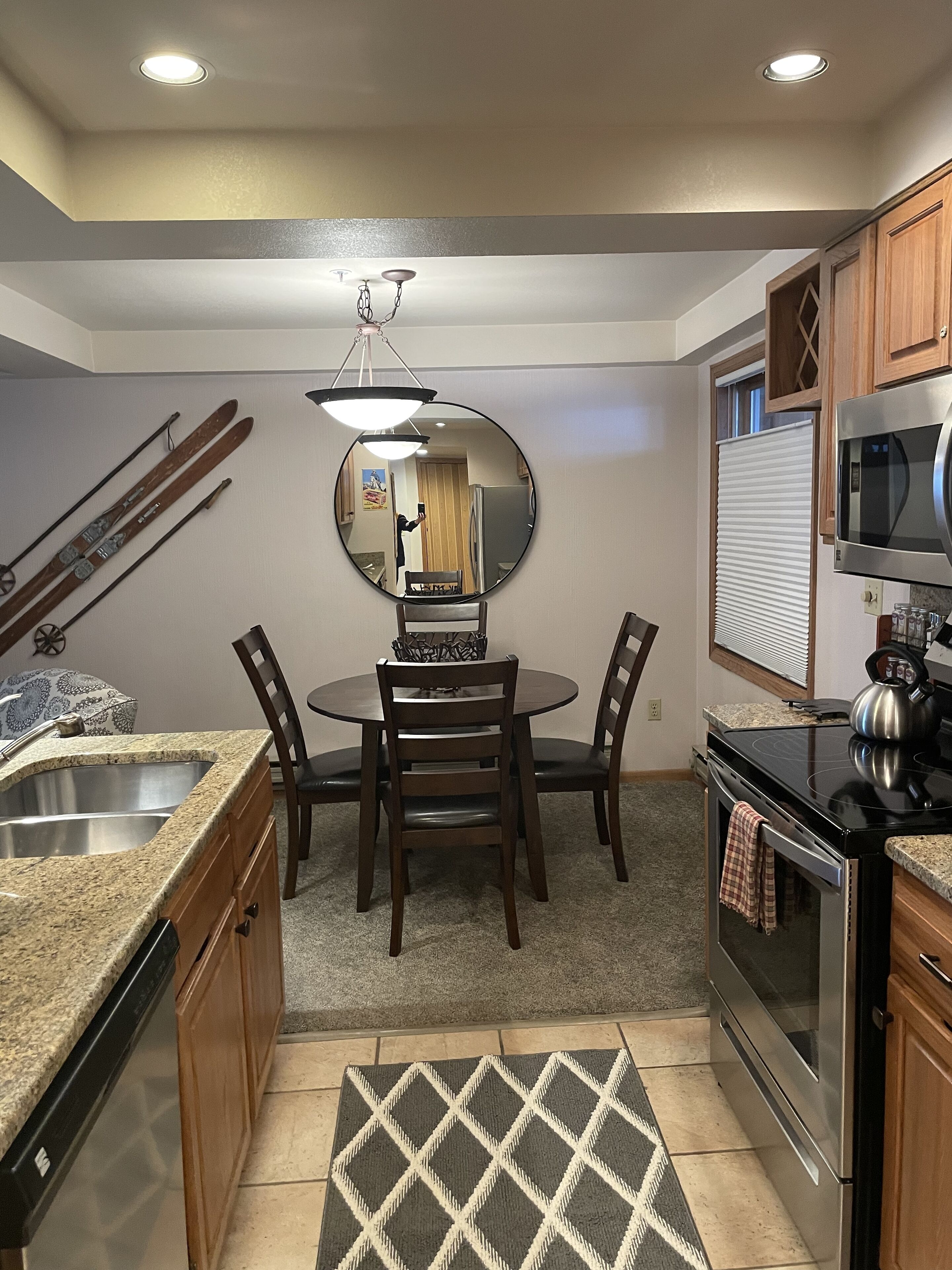 A compact kitchen with a dining table, chairs, a round mirror, a stove, a microwave, and a dishwasher.