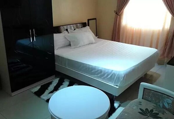 A bedroom with a bed, a round white stool, a wardrobe, and a window with curtains.