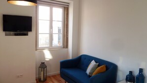 A room with a blue sofa, a TV mounted on the wall, a window with curtains, and a hanging light fixture.