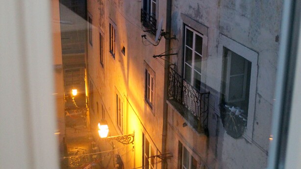 A view through a window of a narrow street with buildings on either side, a bicycle parked outside one building, and a street lamp visible in the distance.