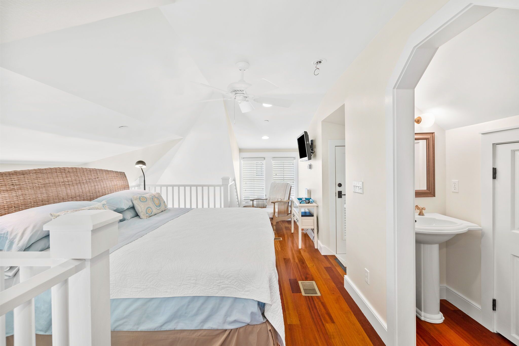 A bedroom with a large bed, a ceiling fan, a white dresser, and a bathroom with a sink and mirror.