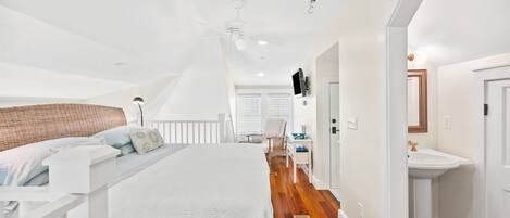A bedroom with a large bed, a ceiling fan, a white dresser, and a bathroom with a sink and mirror.