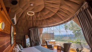 A bedroom with a large bed, a wooden headboard, a small table, and a chair. There is a window with curtains and a view of the outdoors. - Mount Xanadu (Sulthan Bathery)