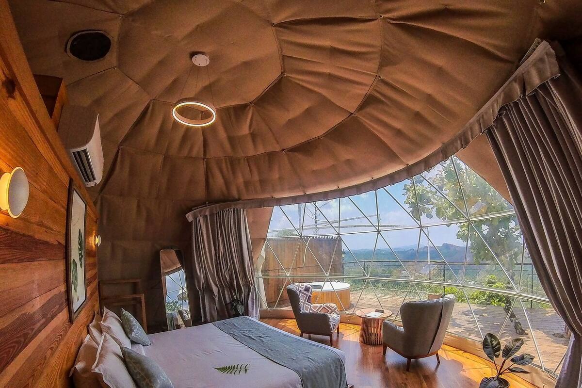 Un dormitorio con una cama grande, cabecera de madera, una mesita y una silla. Hay una ventana con cortinas y vistas al exterior.