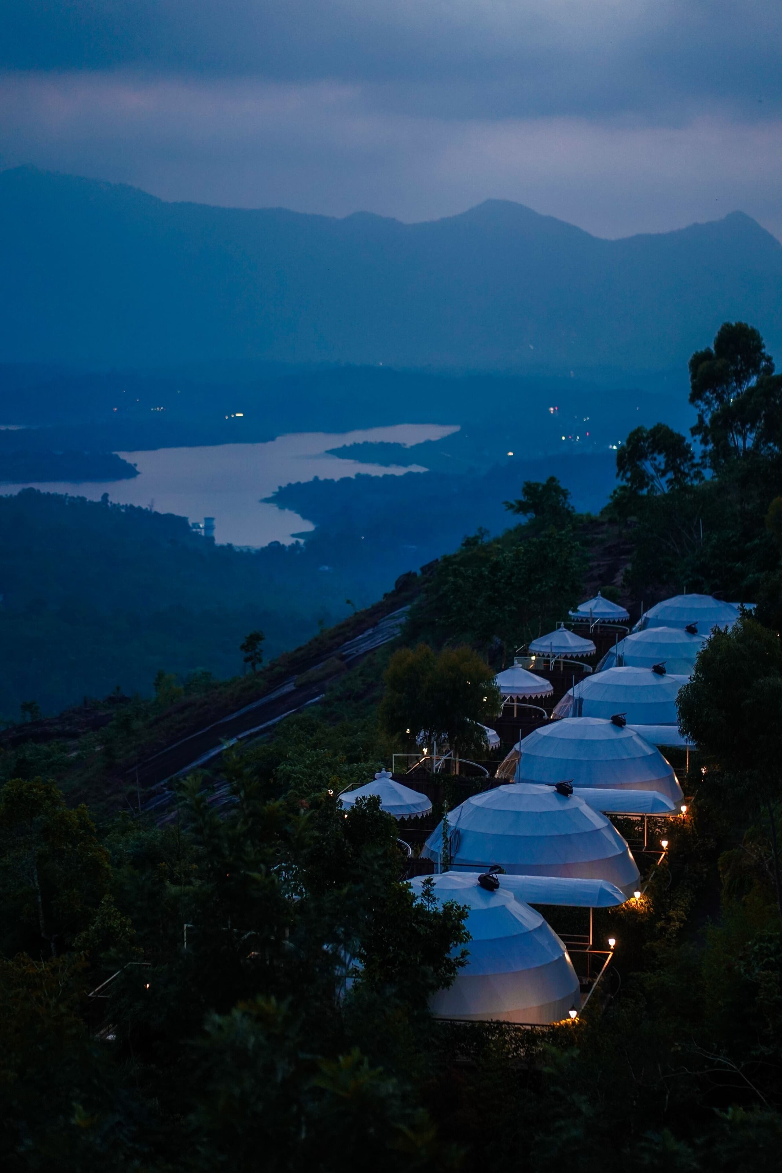 A row of dome-shaped tents with lights, set against a backdrop of mountains and a lake, suggesting a remote, natural setting.