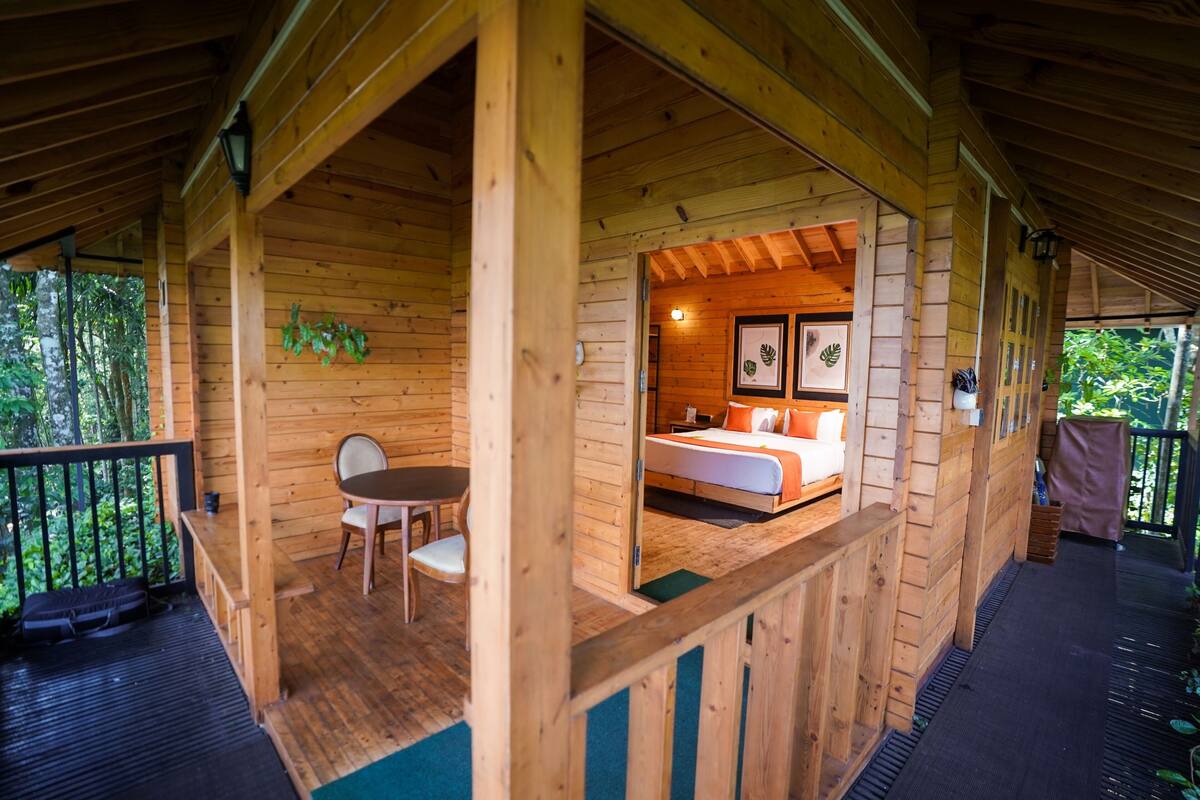 Una cabaña de madera con balcón, una cama con almohadones y una mesa con sillas.