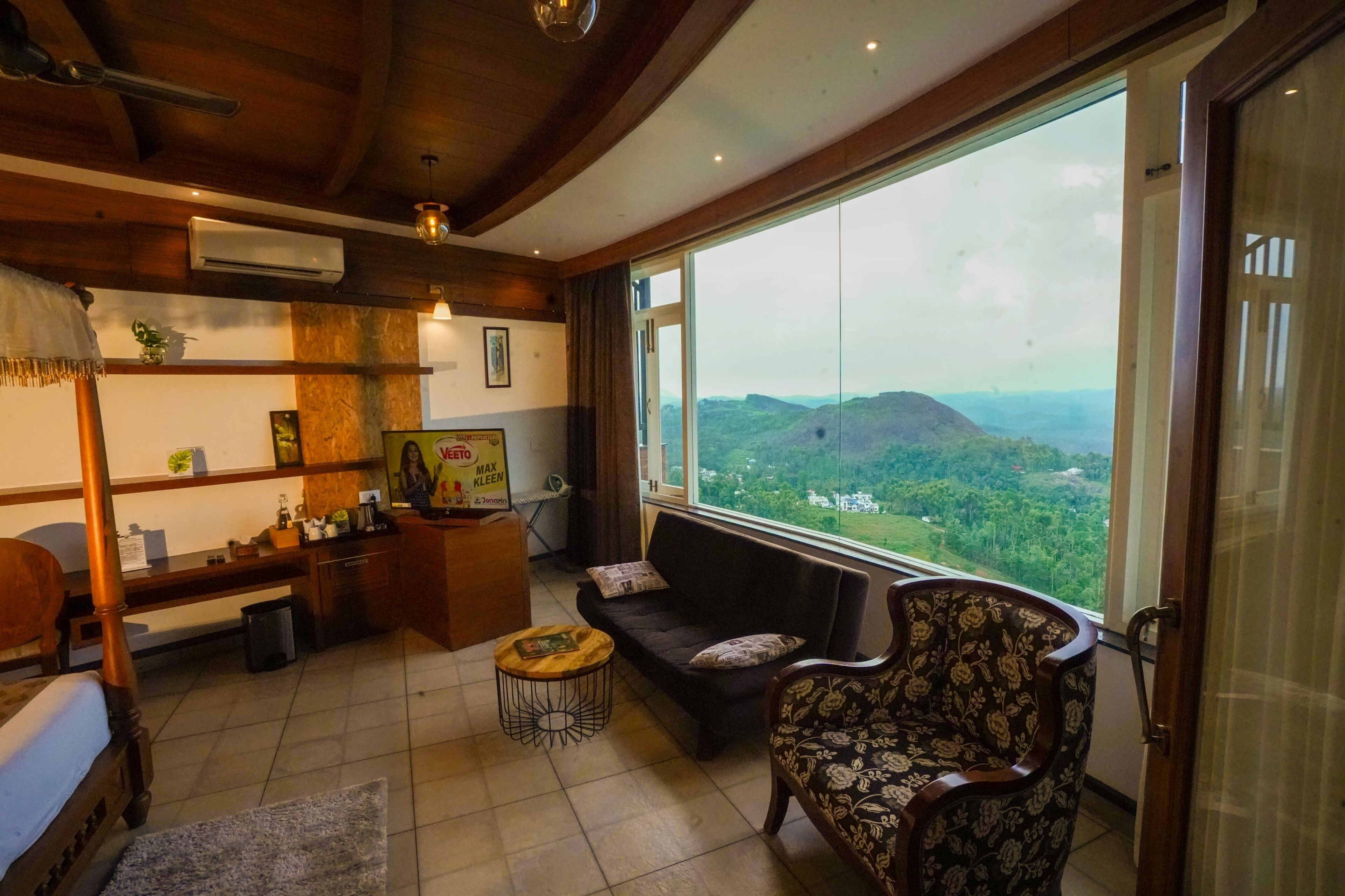 A cozy room with a large window offering a view of the countryside. There is a wooden bed, a sofa, and a patterned armchair.
