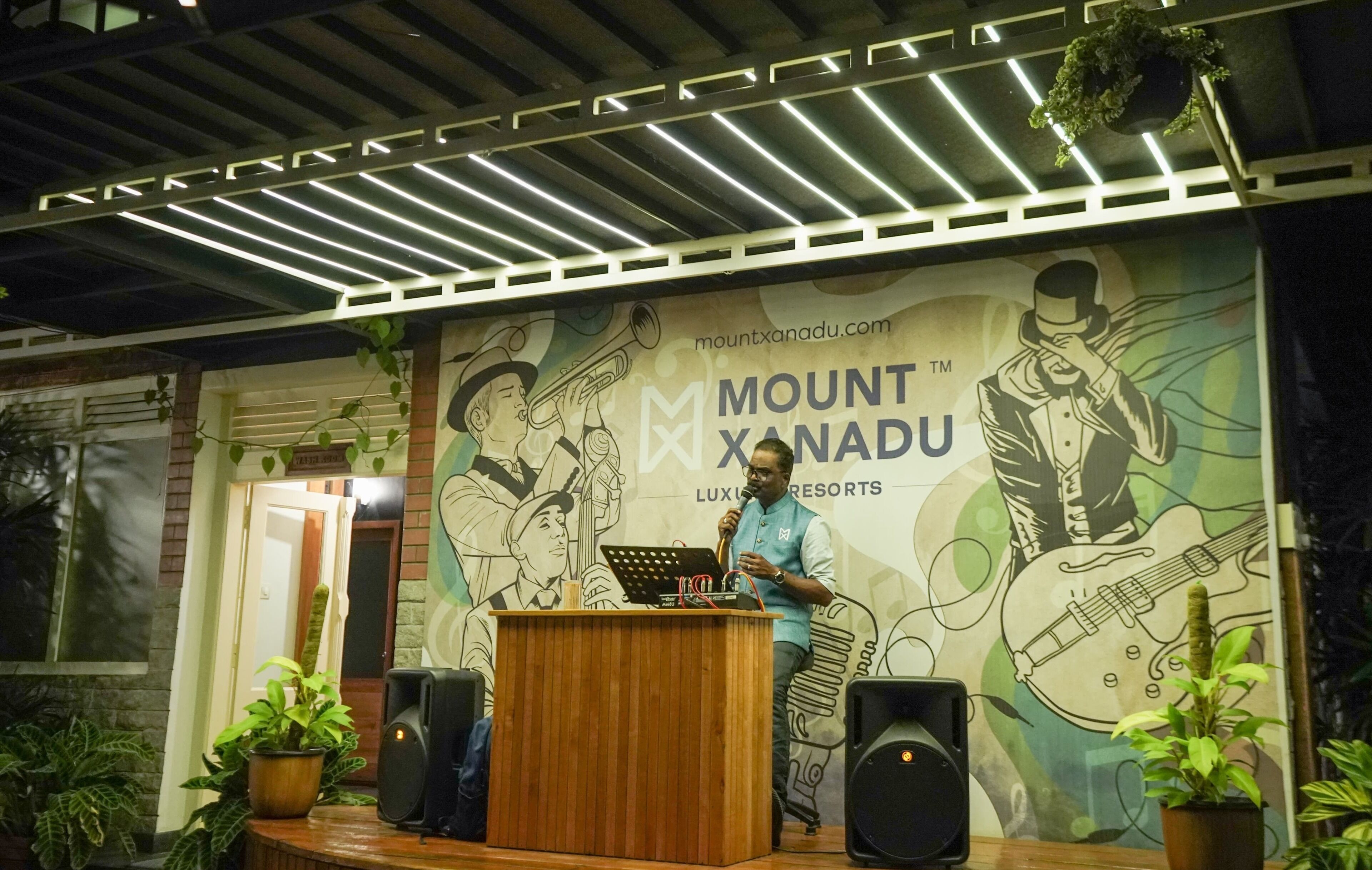 Een podium met een microfoon en een luidspreker, voor een achtergrond met een muurschildering van muzikanten en de tekst 'Mount Xanadu Luxury Resorts'.