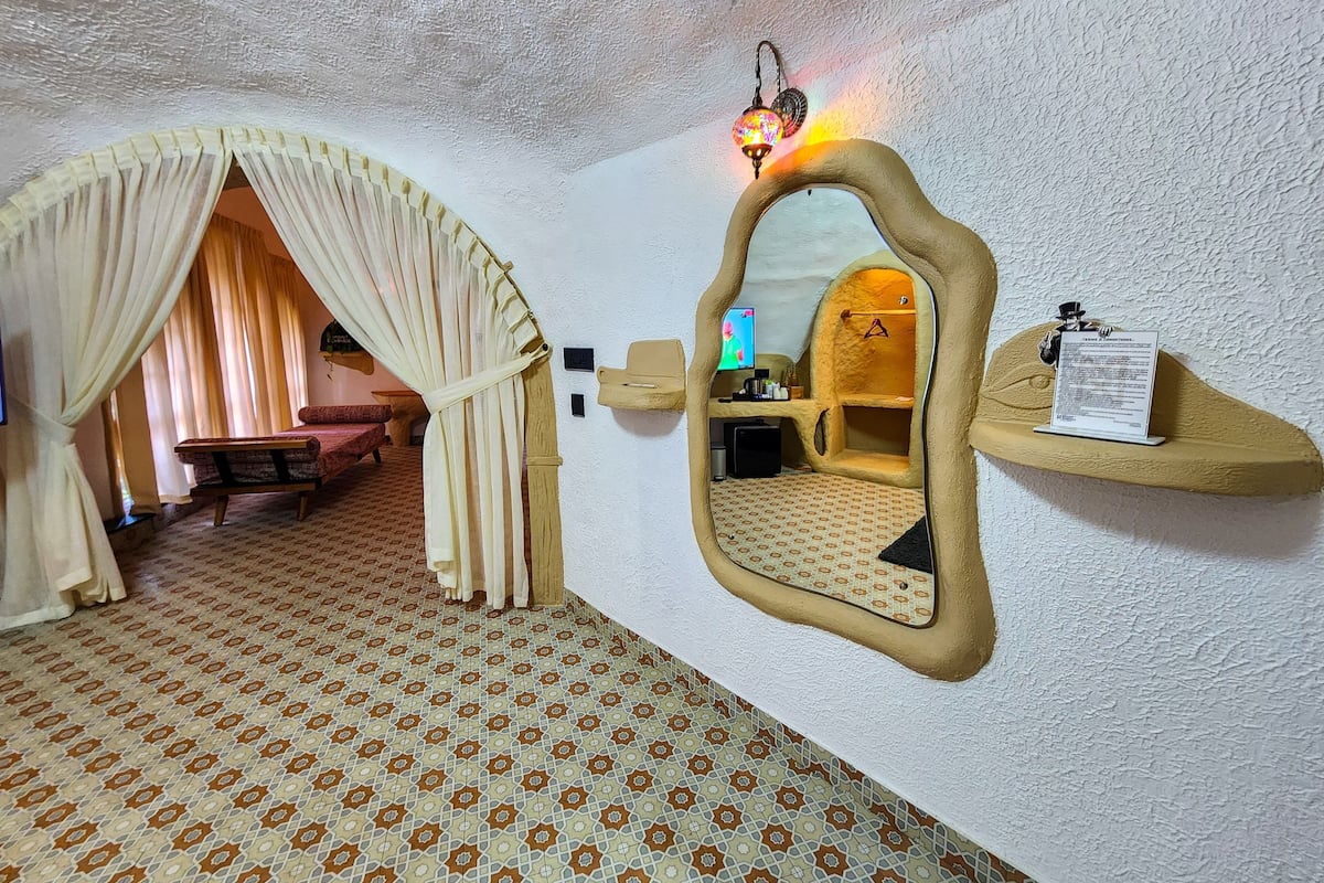 Una habitación acogedora con piso estampado, una cama con cortinas, un espejo, una estantería con una lámpara y un televisor.