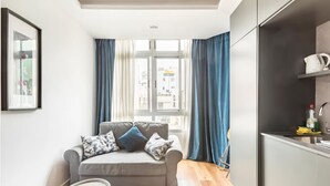 En kompakt opholdsstue med en grå sofa, et lille rundt bord og et tekøkken med elkedel og rengøringsmidler.