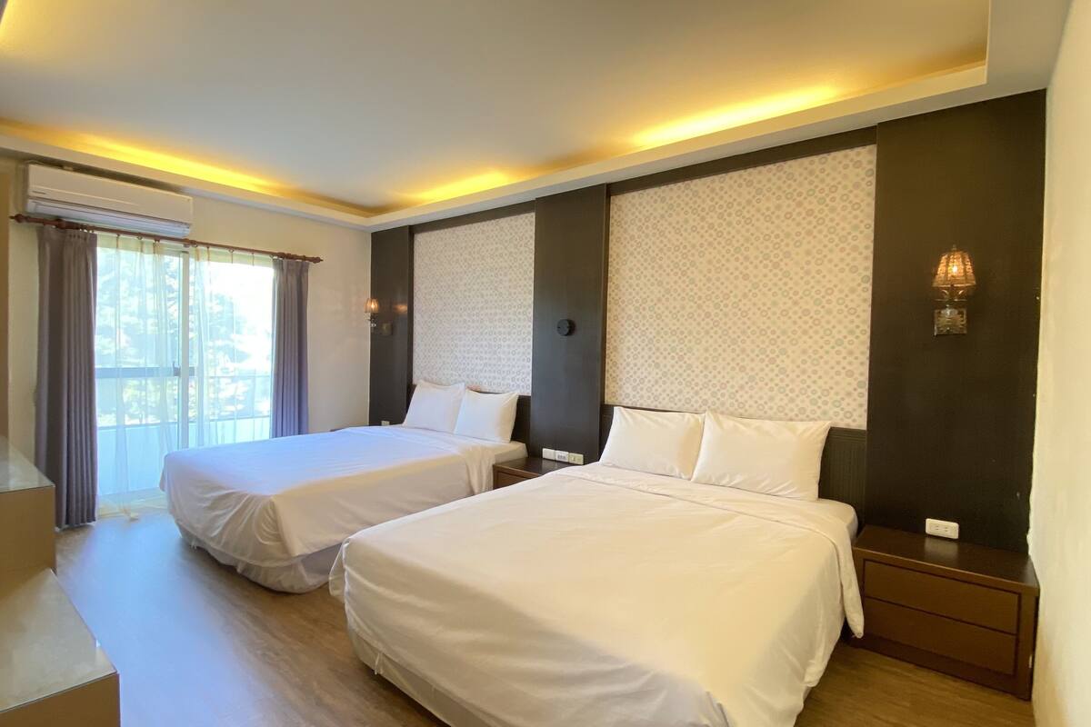 A hotel room with two beds, a large window with curtains, a wall-mounted air conditioner, and a nightstand with a lamp.