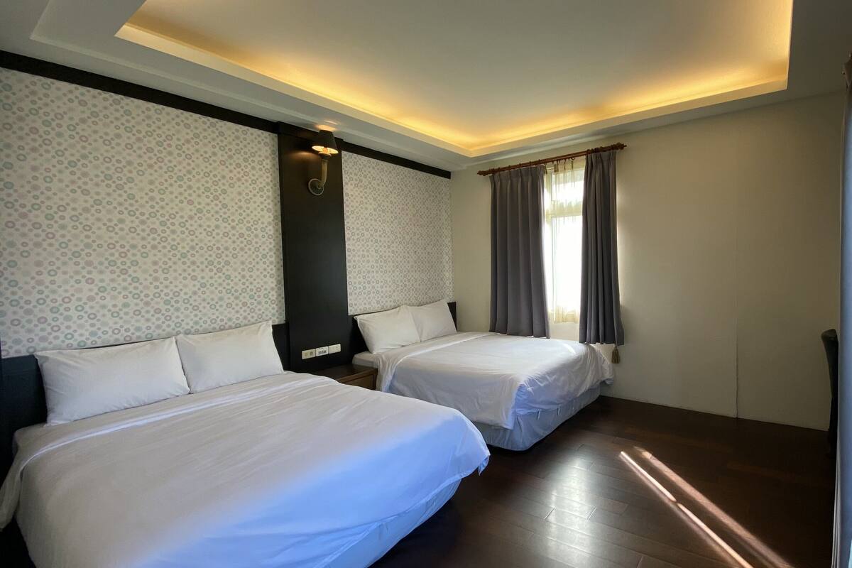 A hotel room with two beds, a nightstand, a wall-mounted lamp, and a window with curtains.