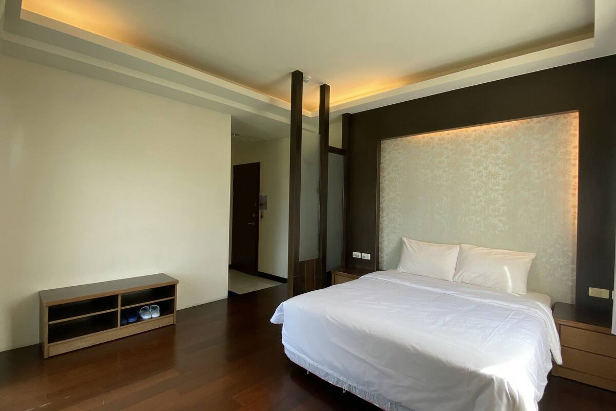 A hotel room with a large bed, a wooden nightstand, a built-in bench with storage, a large mirror, and a door leading to another room.