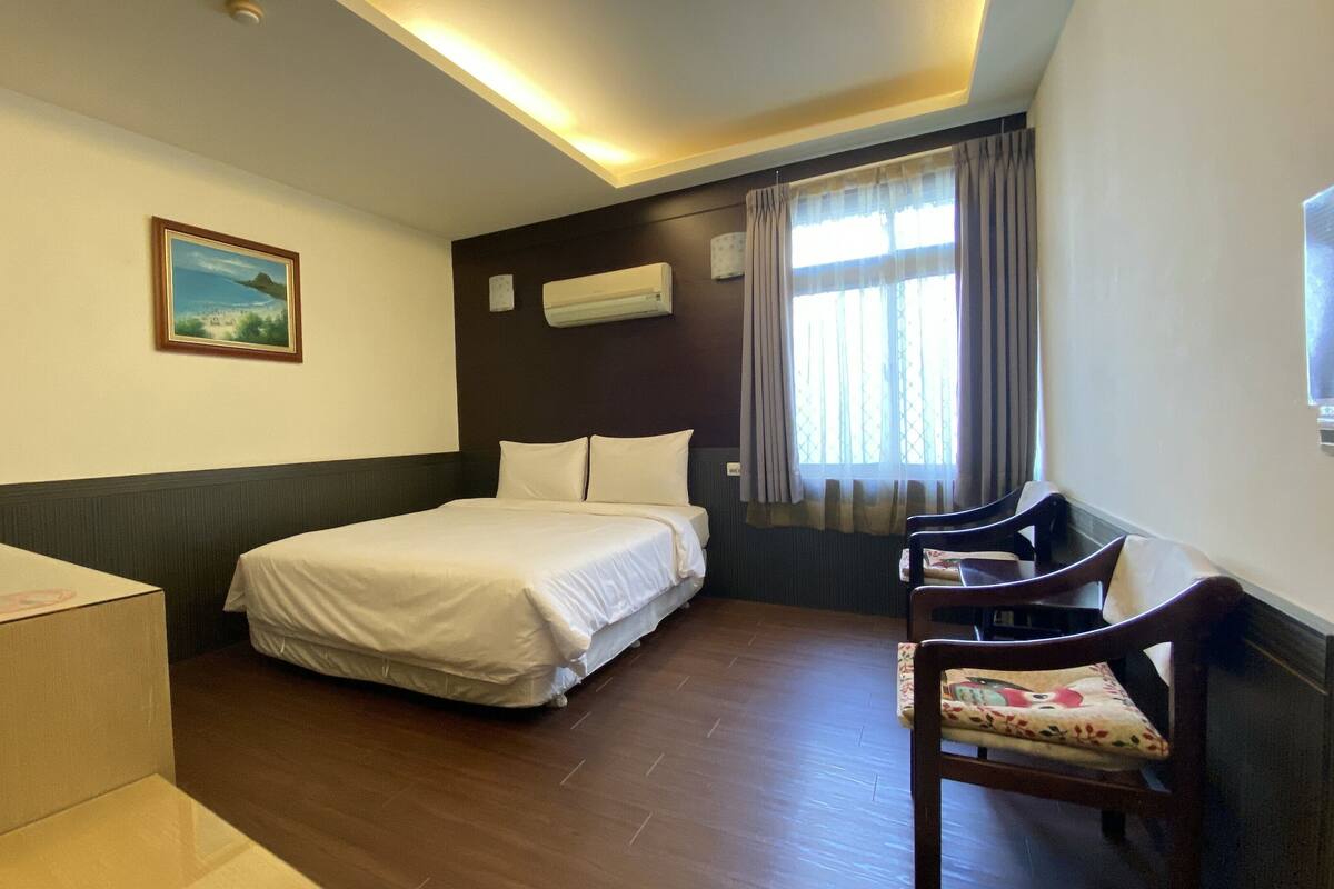 A hotel room with a bed, a desk, a chair, an air conditioner, and a window with curtains.