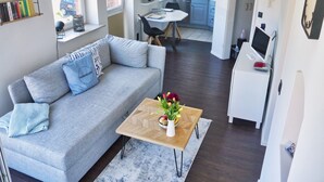 Un salon moderne avec un canapé gris, une table basse en bois ornée d’un vase de fleurs, et une cuisine en arrière-plan.