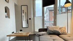 Un salon moderne avec un canapé de couleur sombre, une table basse en bois et une grande fenêtre donnant sur les bâtiments environnants.