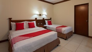 Habitación de hotel con dos camas, cada una con cabecero de madera, ropa de cama blanca y detalles en rojo. Hay luces fijadas a la pared y un ventilador de techo.