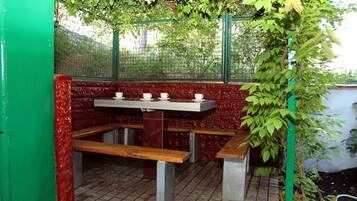 A covered outdoor seating area with a table and benches, surrounded by a green fence and foliage.