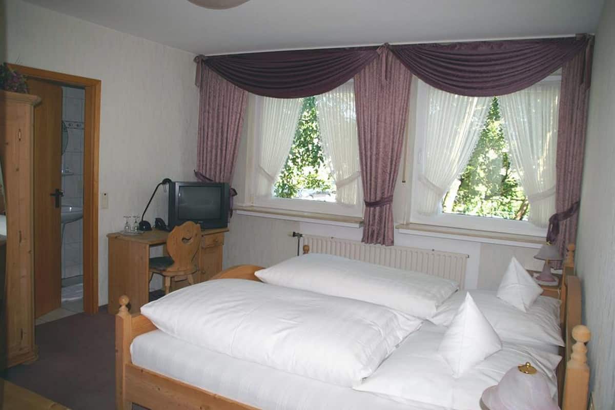 A bedroom with a double bed, a wooden desk with a television, and a window with curtains.