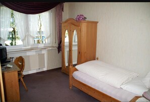 A bedroom with a wooden bed, a wooden dresser, a chair, a desk with a computer, a window with sheer curtains, and a radiator. - Hotel Restaurant Bieberstuben (Menden)